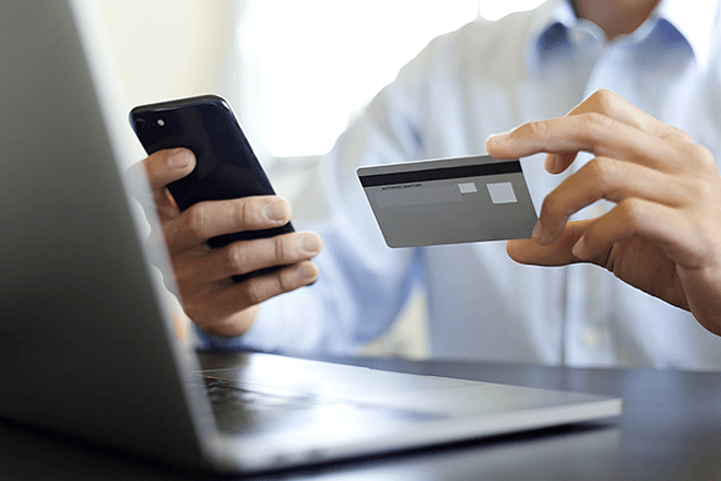 スマートフォンとクレジットカードを手に持ち、ノートパソコンの前にいる人物